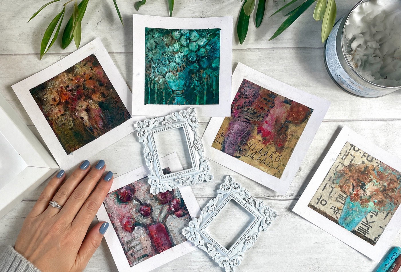 Five small textured flower paintings laid out on white wood with little ornate frames, Marina's hand resting beside them
