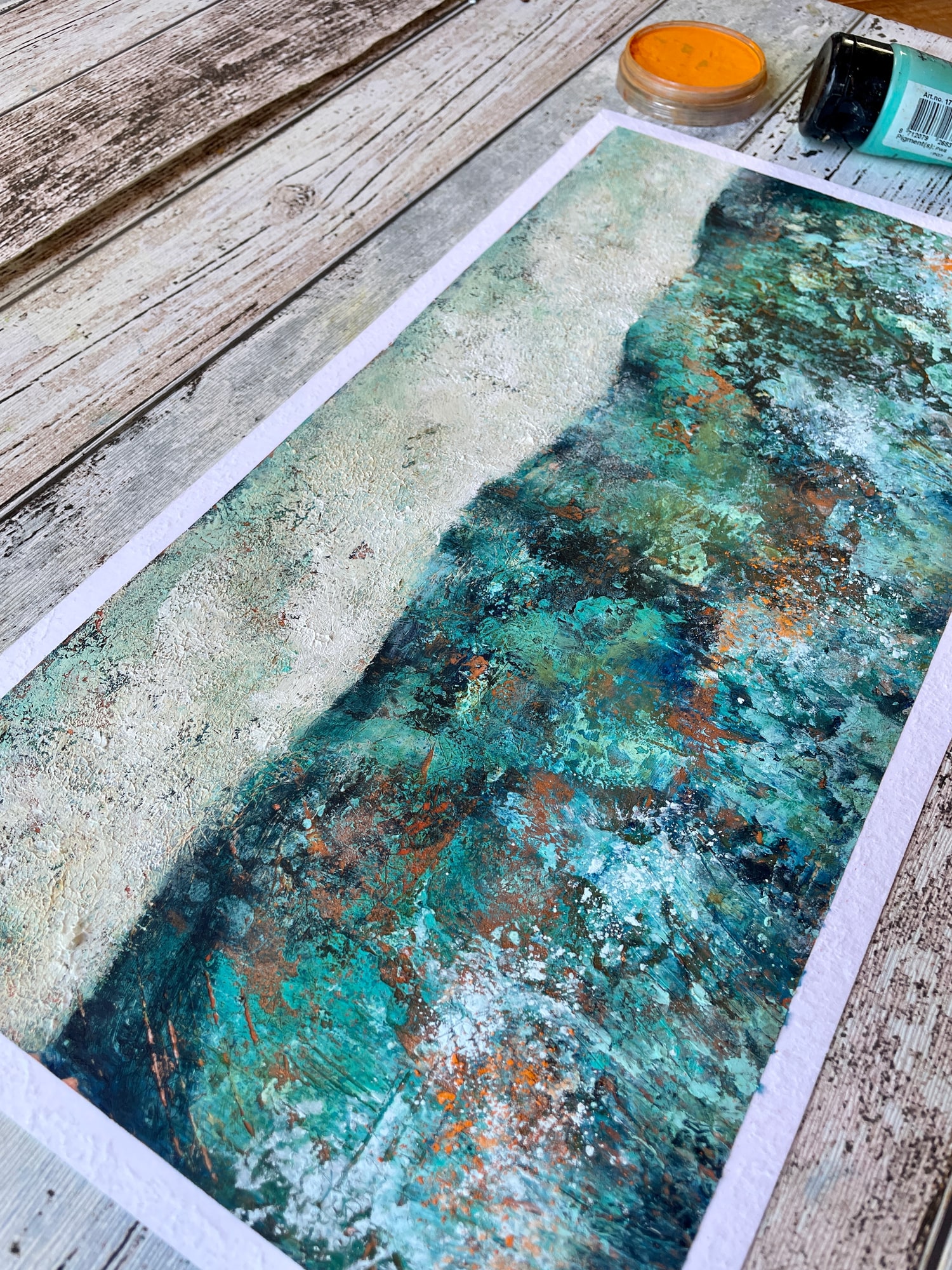 Paysage acrylique turquoise richement texturé sur la table de Marina, avec du pigment orange et de la peinture à côté
