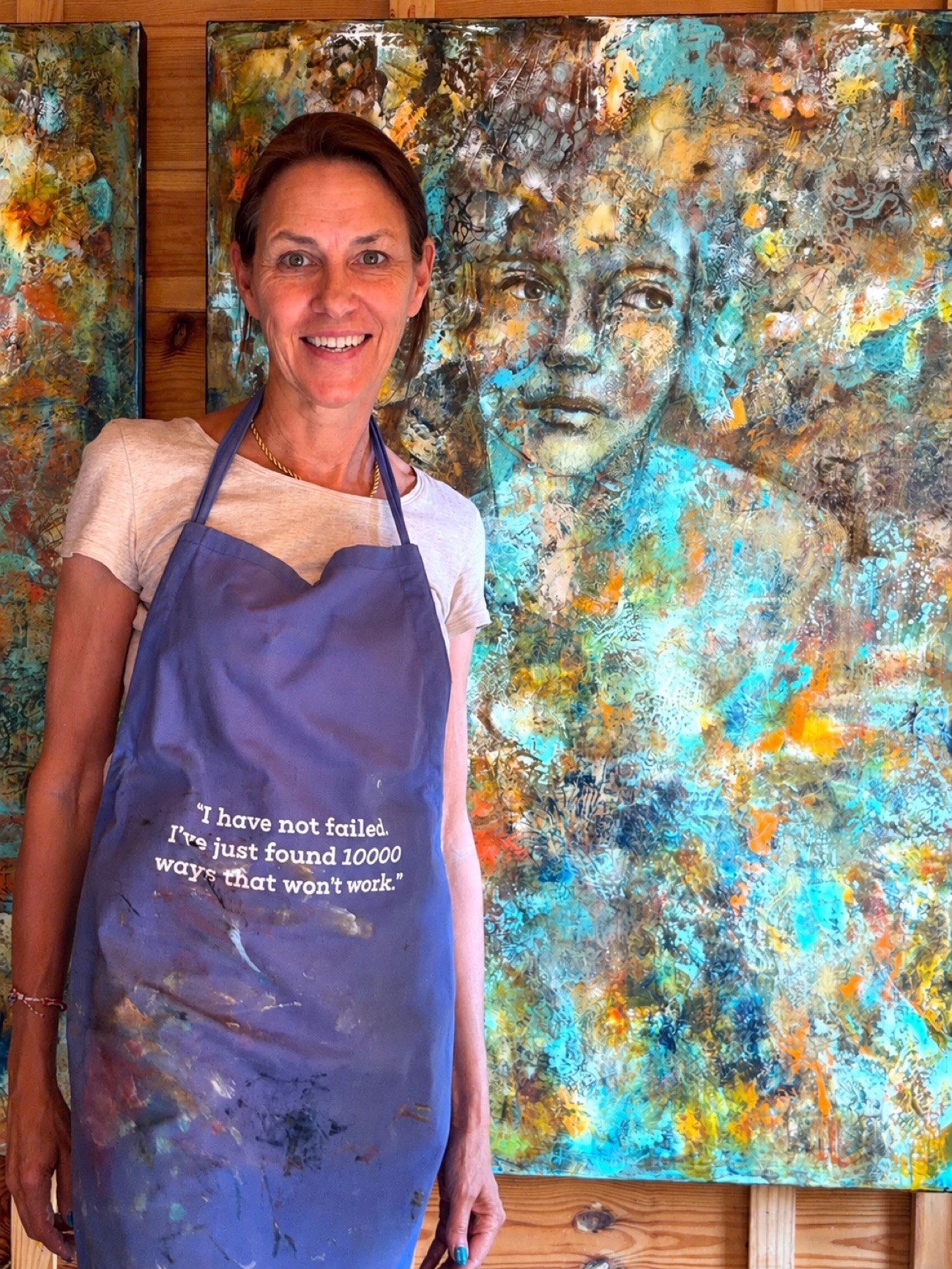 Marina in her painting apron, smiling, in front of a large teal cold wax portrait in her studio